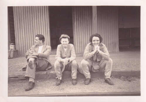 A young man with one shoe off and two young women all sitting on a railway platform.
