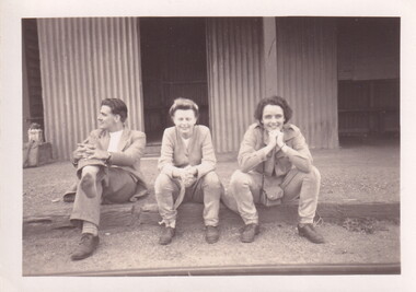 A young man with one shoe off and two young women all sitting on a railway platform.
