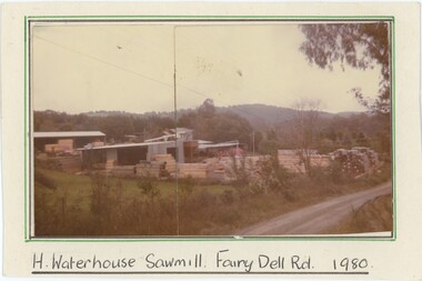 An analogue colour photograph mounted on card, depicting a sawmill with mountains in the background.