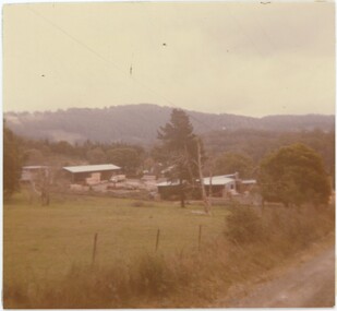 A square analogue colour photograph depicting a sawmill with mountains in the background.