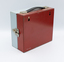 A square metallic box, facing the red rear side with a carry handle at the top and a metal clasp and the blue of the front side visible.