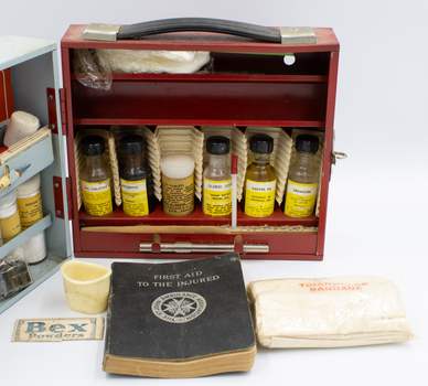 An open metal box with two sections, facing the one painted red. Several items including a first aid guide book, a small plastic cup, a bandage in plastic and a sachet of Bex powder are laid out in front.