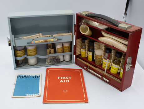 An open metal box with two sections, facing the one painted blue. Two first aid guide books are laid out in front.