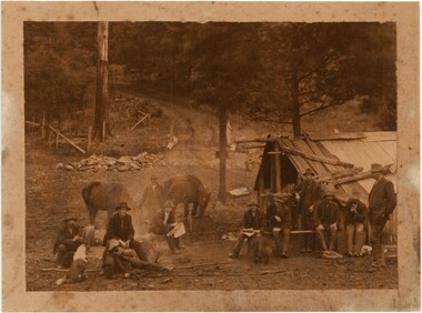 A sepia photograph of a group of men and horses in front of a wooden hut and unpaved road.
