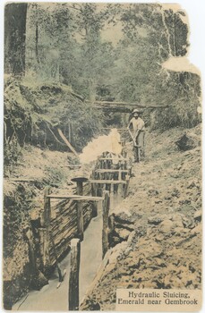 A hand-coloured postcard, caption in bottom-right, showing men besides water flowing through an artificial channel in a forest.