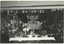Black and white photographic print of a stage decorated with flags, flowers and a large banner.
