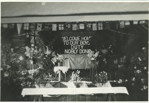 Black and white photographic print of a stage decorated with flags, flowers and a large banner.