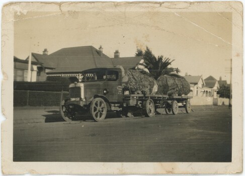 A sepia photograph of a truck pulling trailers with two large logs down a street with homes.