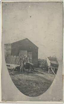 A black and white oval-shaped photograph of a yard with tools, wagons, and a wooden shed.
