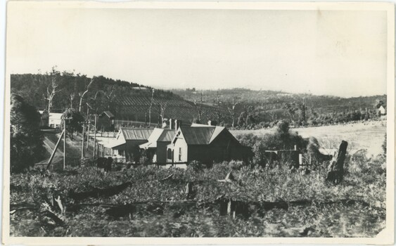 A black-and-white photograph of a house in a field, with hills in the background.