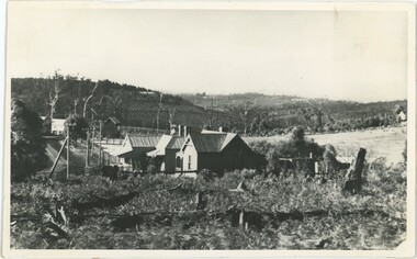 A black-and-white photograph of a house in a field, with hills in the background.