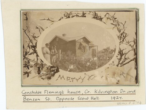 A sepia photograph mounted on card with a handwritten caption below; the photograph depicts a wooden house with two women in front.