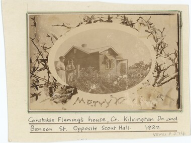 A sepia photograph mounted on card with a handwritten caption below; the photograph depicts a wooden house with two women in front.