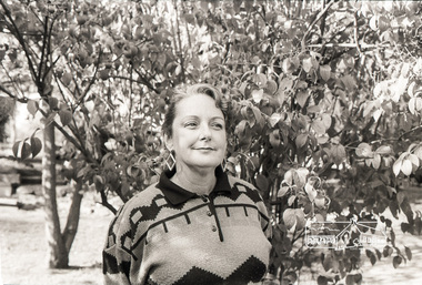 Black and white photo of woman with slick hair and curl on her forehead stands with a closed smile in front of a tree. She is wearng diamond shaped dangly earrings and a knitted jumper with geometric patterns with a black collar and  three buttons in the centre.