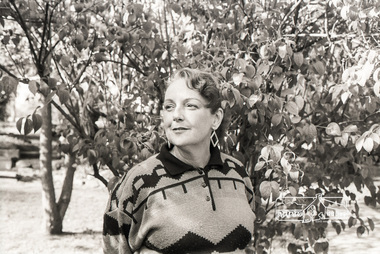 Black and white photo of unknown woman with slick hair and curl on her forehead stands with a closed smile in front of a tree. She is wearng diamond shaped dangly earrings and a knitted jumper with geometric patterns with a black collar and three buttons in the centre.