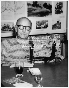 Man in glasss with a hearing device wearing a woolen jumper seated at a table holding up a hard cover book with the title "Pioneers & painters: One hundered years of Eltham and its Shire" Alan Marshall.