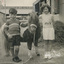 Black and White photo of a young boy and slightl olfer girl wearing knitted jumpers with a pony.  There is a rope lead on the pony.  A pile of hay int he background and a sign on a building in the background reading "dogg'em cars'.