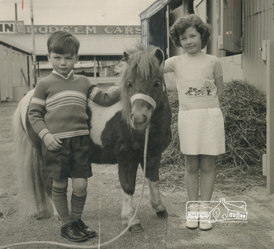 Black and White photo of a young boy and slightl olfer girl wearing knitted jumpers with a pony.  There is a rope lead on the pony.  A pile of hay int he background and a sign on a building in the background reading "dogg'em cars'.