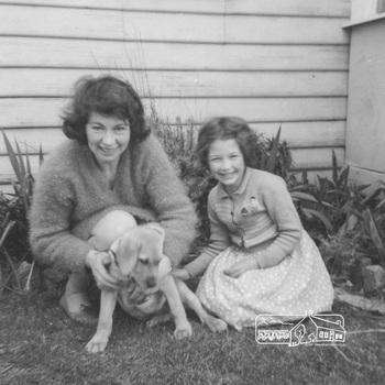 A woman is partl kneeling on the grass holding a puppy.  A young girl wearing a dress and knitted jumper is sitting on the grass touching the rear of the puppy.  There is a weatherboard builidng in the background with a grden bed betweeb them.  Ada and Susan are smiling and looking directly at the camera