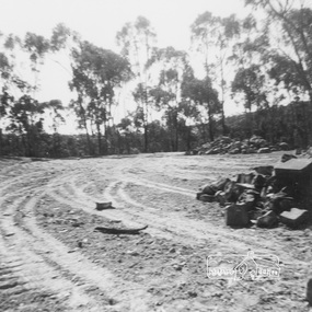 An expanse of dirt with wheel tracks, with trees and bush in the background and piles of rocks on the right hand side