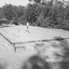 A boy with a bow and arrow stands on a flat clear block of land where straight pieces of timber have been liad out to form the shape of a house