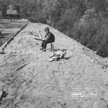 A woman sits on a temporary wooden structure at the edge of the cleared flat block of land close to the concrete base of the house
