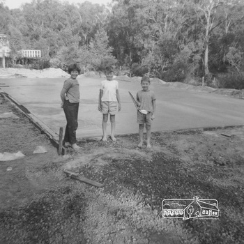 A young girl and two boys stand in front of a large sqaure of concrete on a flat block of land