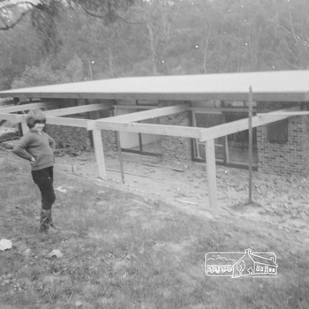 New brick house with windows, flat roof and outdoor pagola under construction.  A young girl stands in the foreground on a hilly grassed verge with her hands on her hips