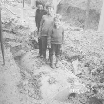 A young girl and two boys stand in a row on top of a buried concrete tank amid building rubble beside their newly constructed house and underneath the pagola