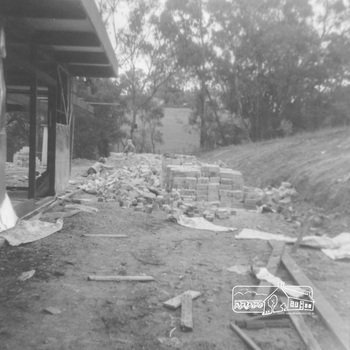 View of side of house under construction with stacked bricks and brick and building rubble. Children play on a dirt pile n the background with bushline in the distance