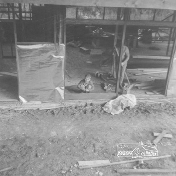 A young child crouches on the floor beside an adult who is sweeping the concrete floor.  He is looking out to the photographer past an area of flat hard dirt at the side of the house There is a roof, but no walls.