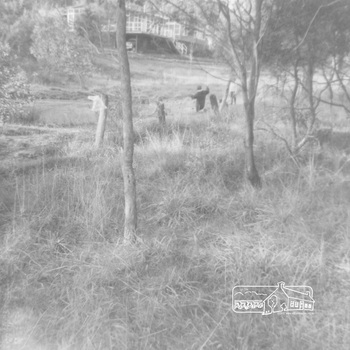 A partial view of a house in the distance with prominant white framing.  A dam is in the middle of the photo with two children standing near it.  The block has long grass and  sapling trees growing in the foreground.  A  rugged wire fence  runs along the middle of the photo