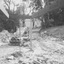 A woman and young boy near a VQ Beetle paked in front of piles of dirt and brick rubble and pipes.  A sloped tree lined Metery Road is in the background 