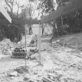 A woman and young boy near a VQ Beetle paked in front of piles of dirt and brick rubble and pipes.  A sloped tree lined Metery Road is in the background 