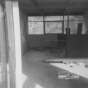 View of room with concrete floor under construction.  There is a ceiling and side wall.  A large shadow falls on the floor.  Pieces of timber are piled on the floor.  The opposite side has big windows looking towards bushland
