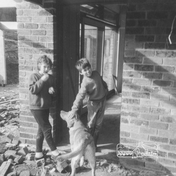 Girl and boy stand at an external door way of a brick house, near a corner.  The boy pats a dog who is wagging its tail with its rear view to the camera.  Brick rubble surrounds the exterior