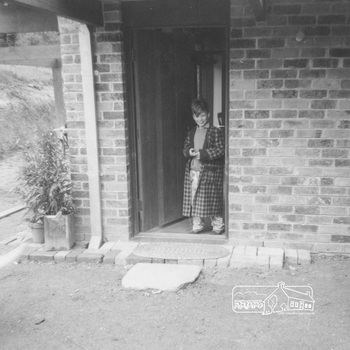 A young boy dressed in a dressing gown smiles at the camera.  He stands inside a brick house with the door open.  The entry has brickwork and mats outside the door.  Plants on containers are sitatuted at the corner of the house