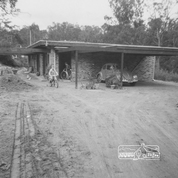 A large unclutterd dirt driveway leads to the brick house and car port with VW Beetles reverse parked.  Two boys ride two-wheeler bikes