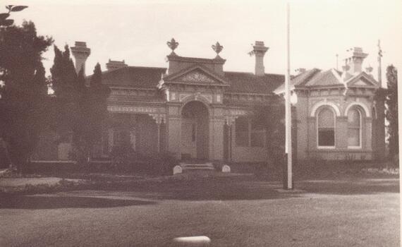 Black and white photograph of the front of an old Victorian-era mansion with features including chimneys, verandah and flag pole.