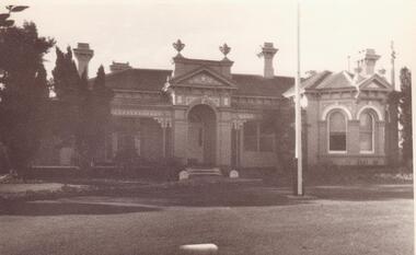 Black and white photograph of the front of an old Victorian-era mansion with features including chimneys, verandah and flag pole.