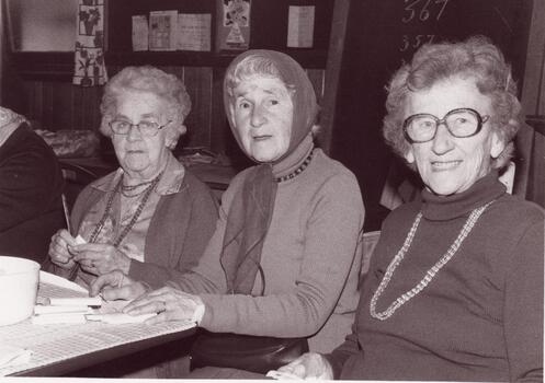 Three older women sitting side by side at a table, indoors.
