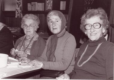 Three older women sitting side by side at a table, indoors.