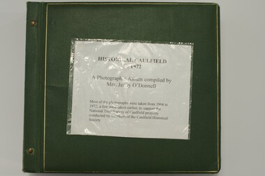 Large book of photographs with a hard, green cover which has 2 bolts holding the pages and cover together.