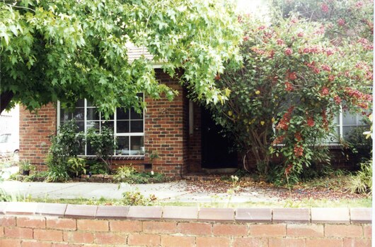 Partial view of the front of a mixed brown brick house in its treed garden behind its unpainted brick fence
