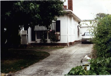 Partial view of white house with brick features, with black and white window awnings and tall chimneys, beside its driveway with an old car in front of the garage.  Garden includes a big tree and smaller plants, with a letter box labelled 4 amongst the greenery at the front.