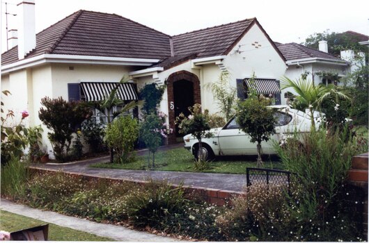 White house with brick features, tall chimneys, striped black and white awnings and arched entry to front porch, in its garden with low brick side fence and concrete drive along the fence line.  There is a car on the lawn amidst the plants.