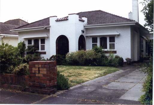 Full front view of white house with brick features, central fort-like porch with 2 doors, 2 tall chimneys, brown brick fence, front garden and concrete drive to the right.