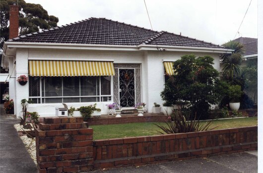 Full front view of white rendered house with brown brick features including chimney, garden bed walls and front fence.  Old car visible in garage up the drive to the left of the house and garden.