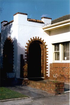 Fortress-like central white entrance porch with 2 open brown brick arches with steps, decorative glass in the top left sash window and a brown brick low wall over the concrete path.