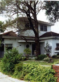 Partial front view of 2 storey white house, weatherboard for the top level and rendered ground floor, with a porch, porthole window by the door and striped awnings.  Large tree in the garden bed amidst smaller plants in front of the porch, with an ivy-covered low side fence with brick pillar.